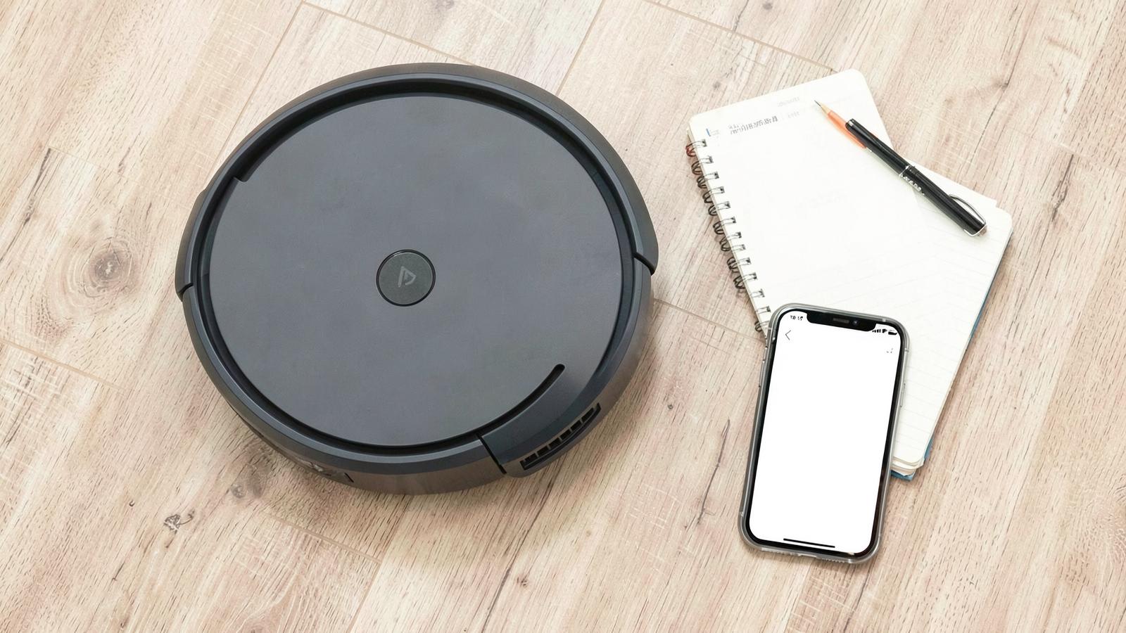 Robot vacuum on a light floor beside a notebook and phone while researching